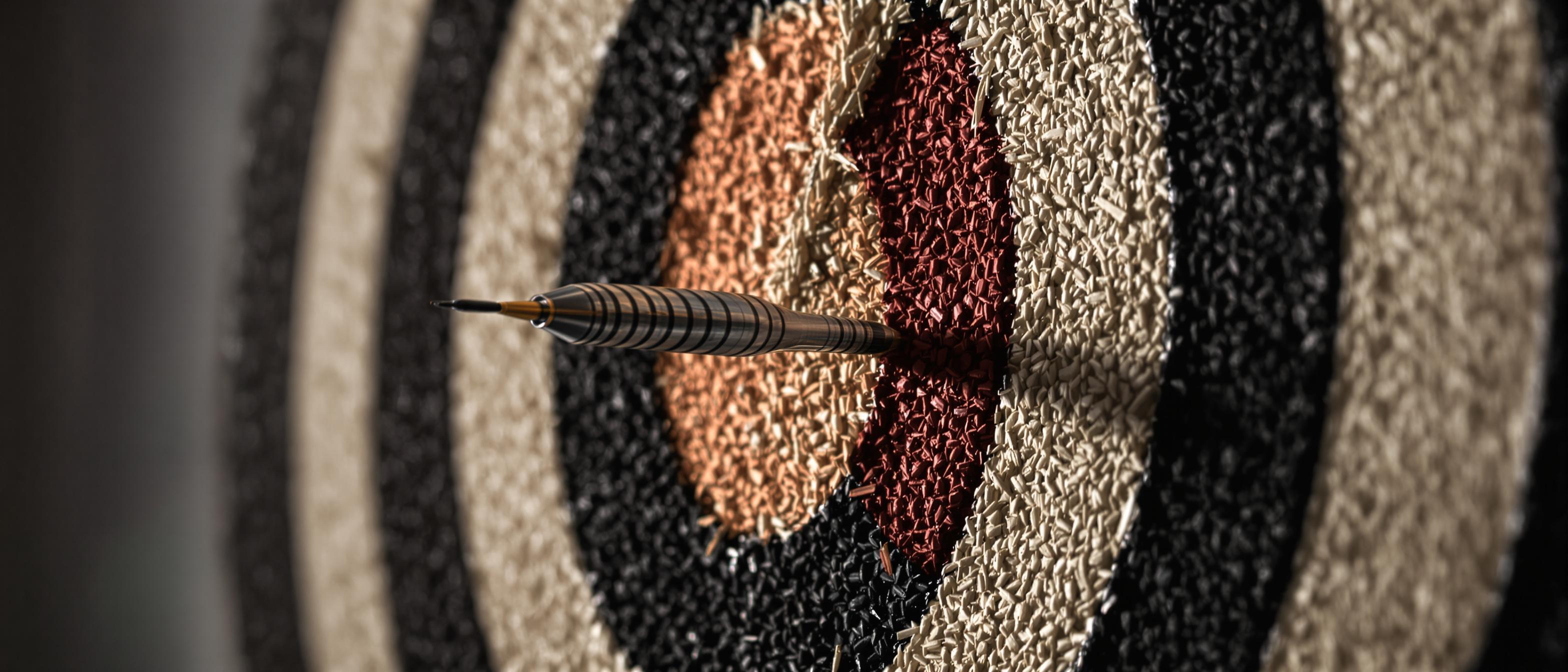 A dart pierces the center of a weathered dartboard in a close-up shot.