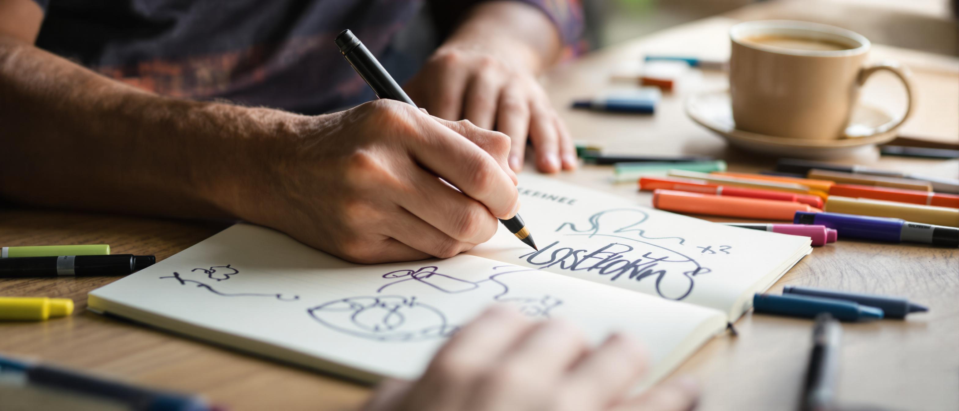 Close-up of hands sketching in a notebook surrounded by art supplies, emphasizing the creative process.
