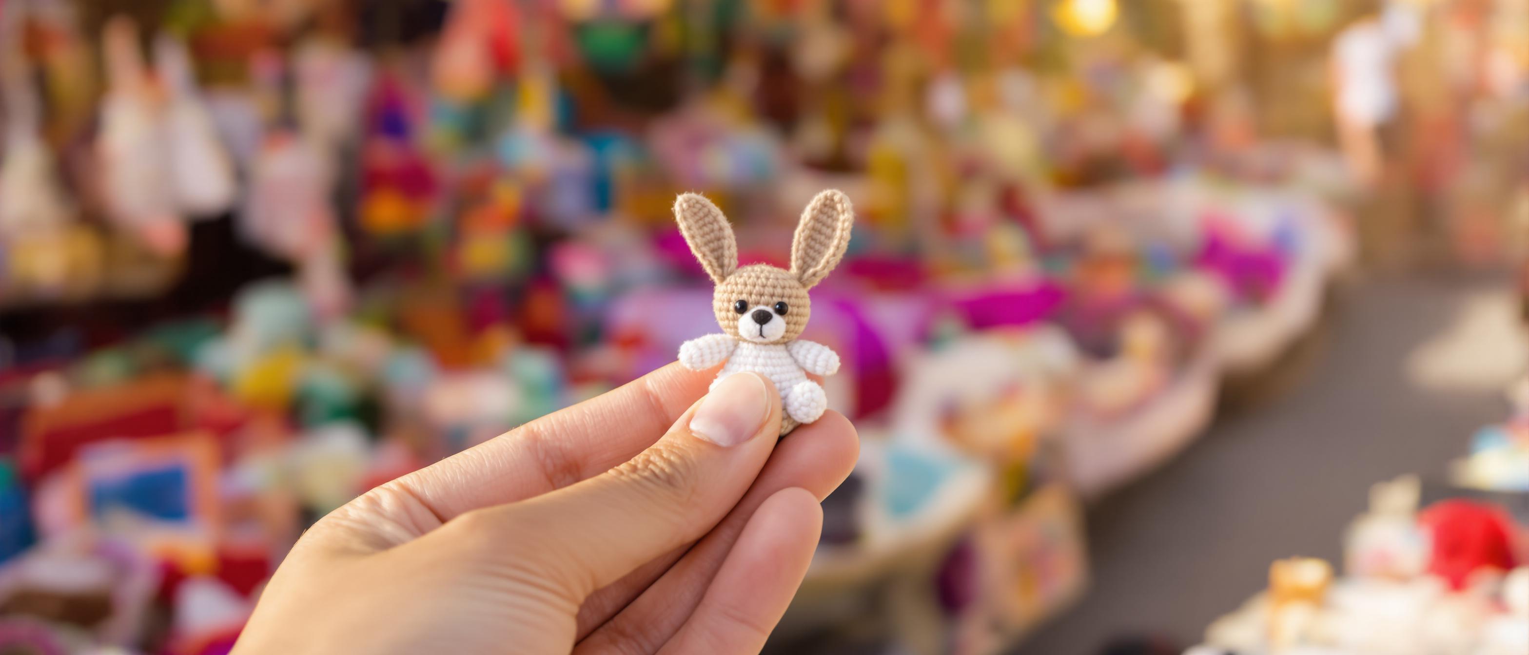 A close-up photo features a hand delicately holding a small, handcrafted item against a blurred background of vibrant crafts.