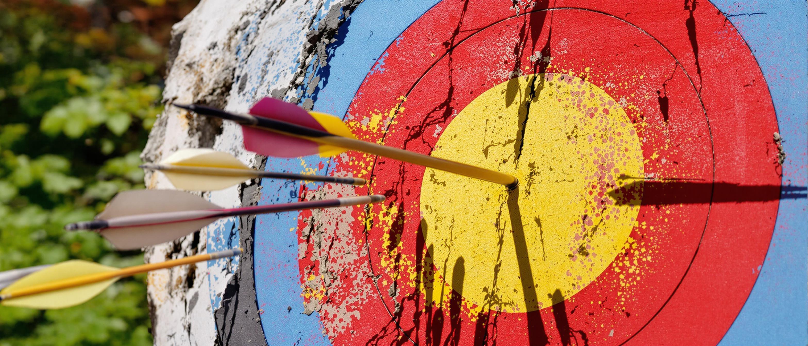 An arrow strikes the bullseye of a classic archery target, with scattered arrows nearby indicating previous attempts.