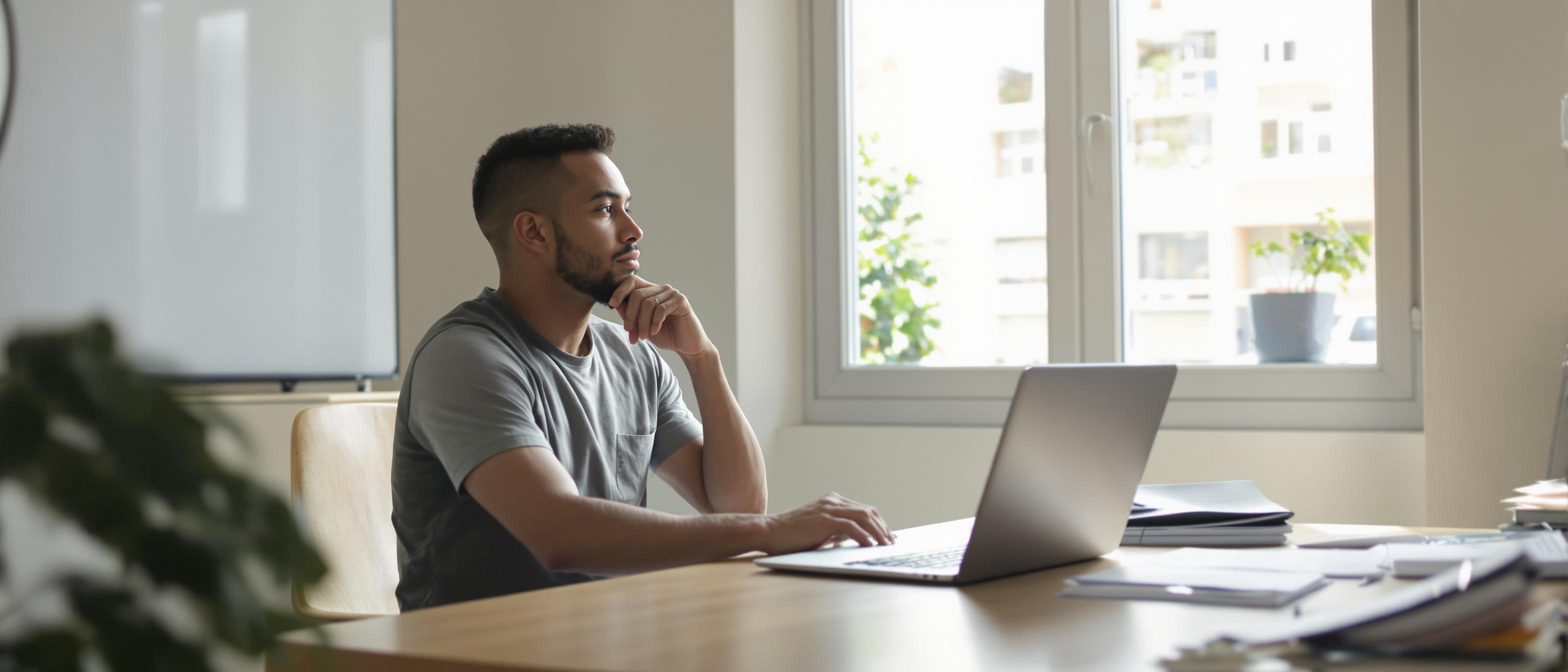 A person sits at a desk, gazing out a window in a brightly lit home office, deep in thought.