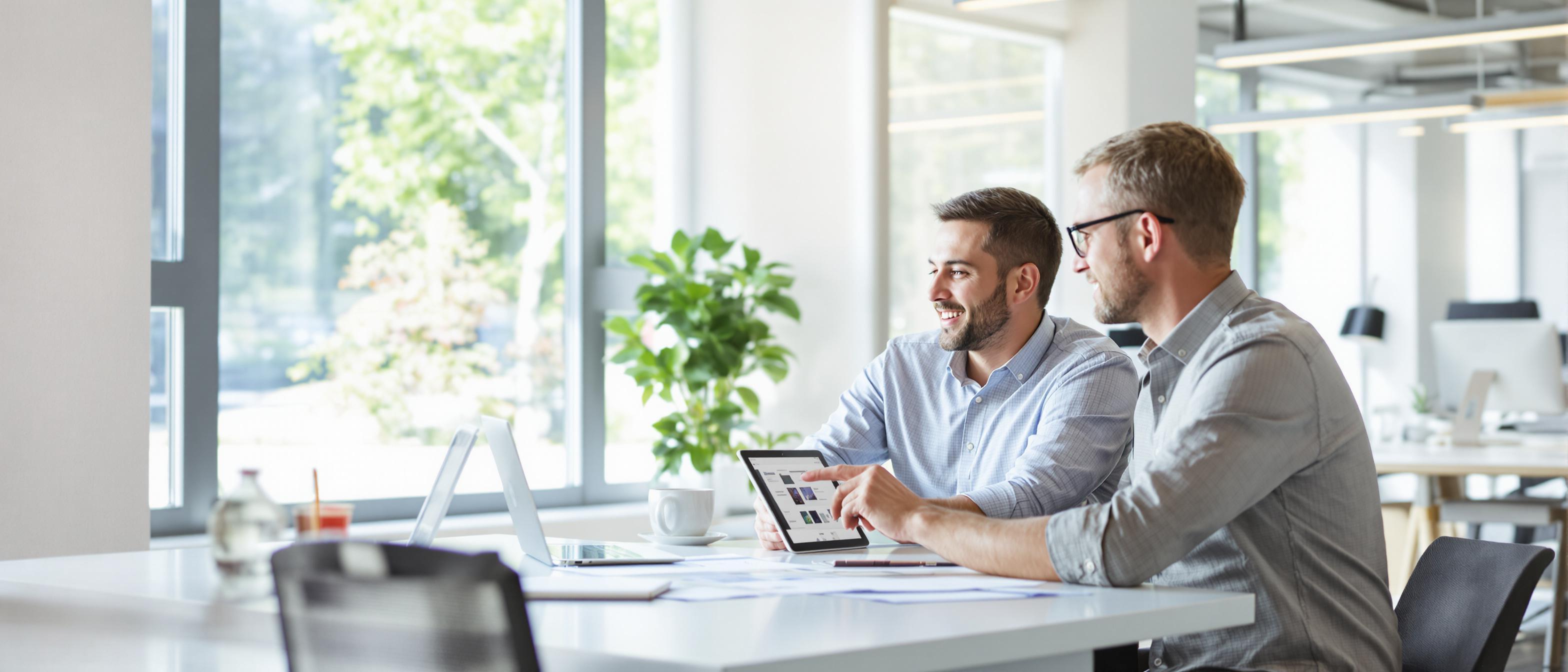 Two individuals engaged in a collaborative discussion while reviewing designs on a tablet in a well-lit office setting.